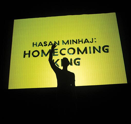 Hasan Minhaj waves in front of a screen saying "Homecoming King"