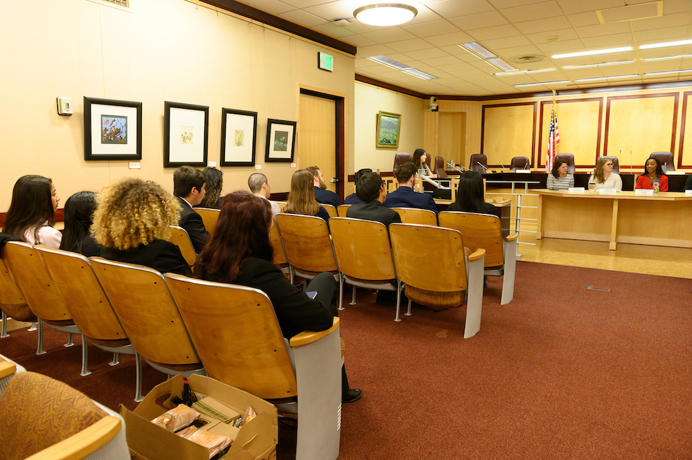 Students sit in a town hall with a podium at the State Capitol Building. 