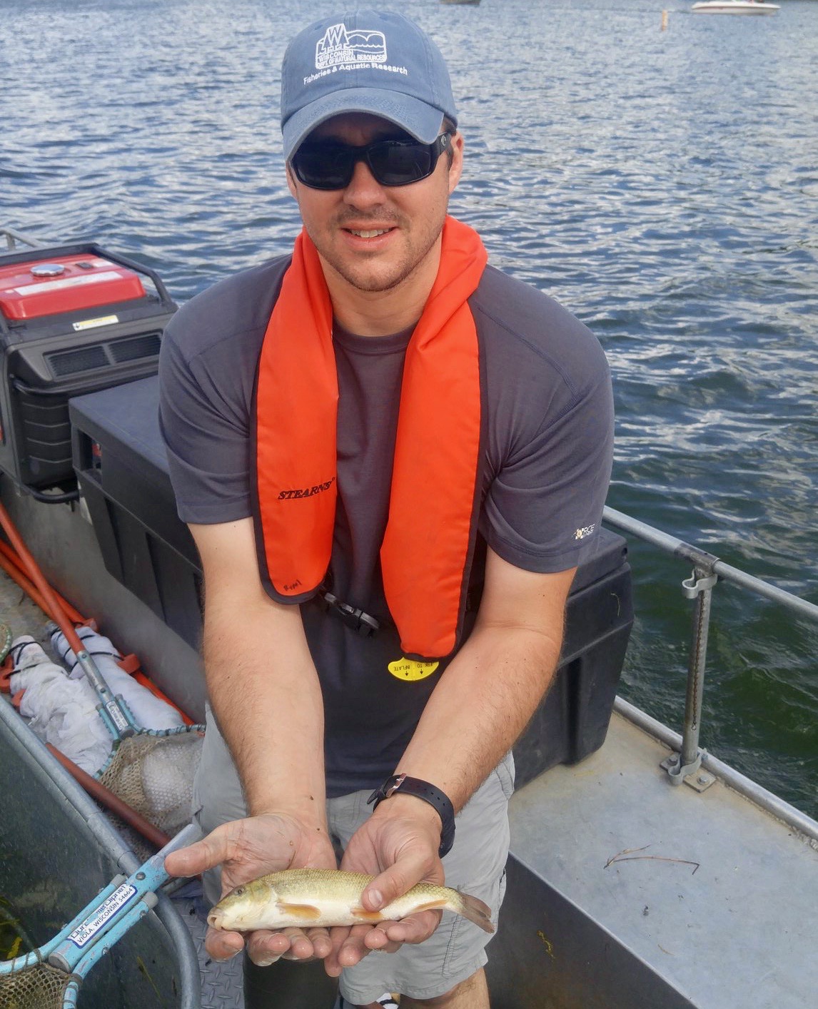 Man in blue ballcap and orange life vest sits on boat holding a sucker fish with lake in background