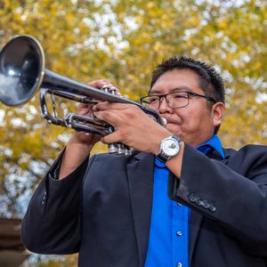 Man plays trumpet with tree behind him