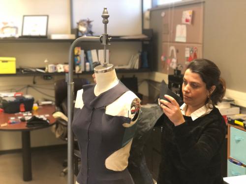 A woman in a black top with brown hair tied back works with a dressmaker's dummy in a lab setting. 