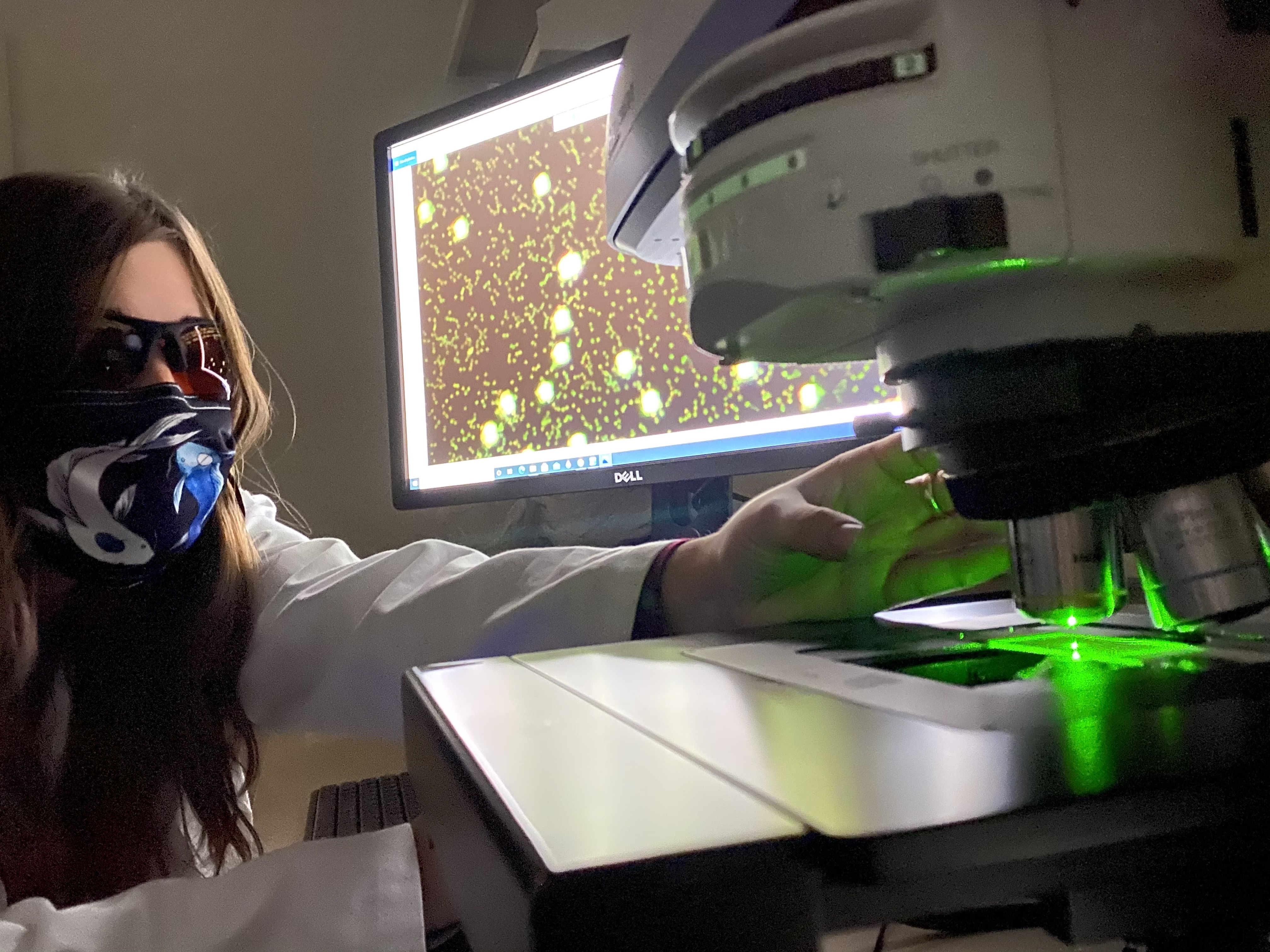 A woman scientist in mask at computer and scientific instrument with green laser