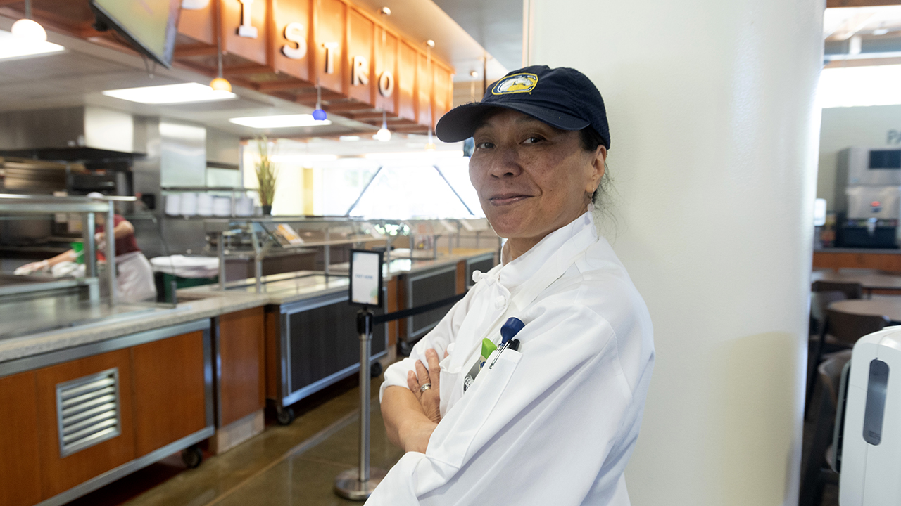 A woman in kitchen whites is photographed in front of the Bistro serving platform at Segundo Dining Commons