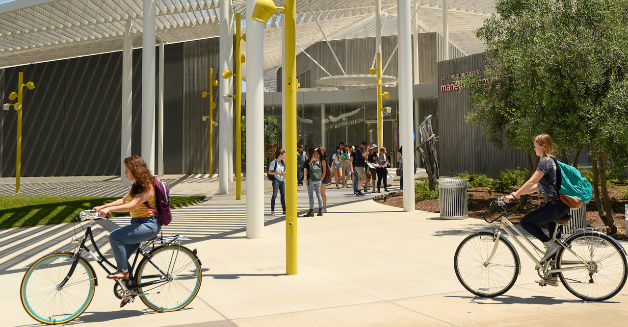 two students riding bikes in front of the Manetti Shrem museum