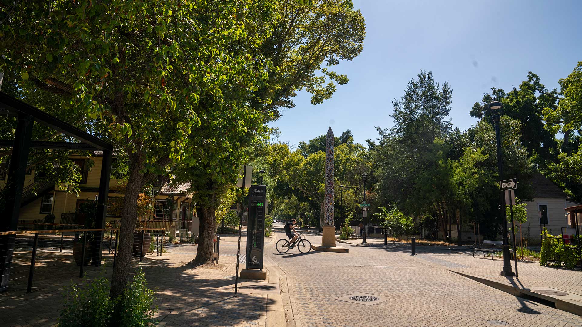 Photo of a tree lined street with an obelisk in the middle of it. A bicyclist rides past the obelisk.