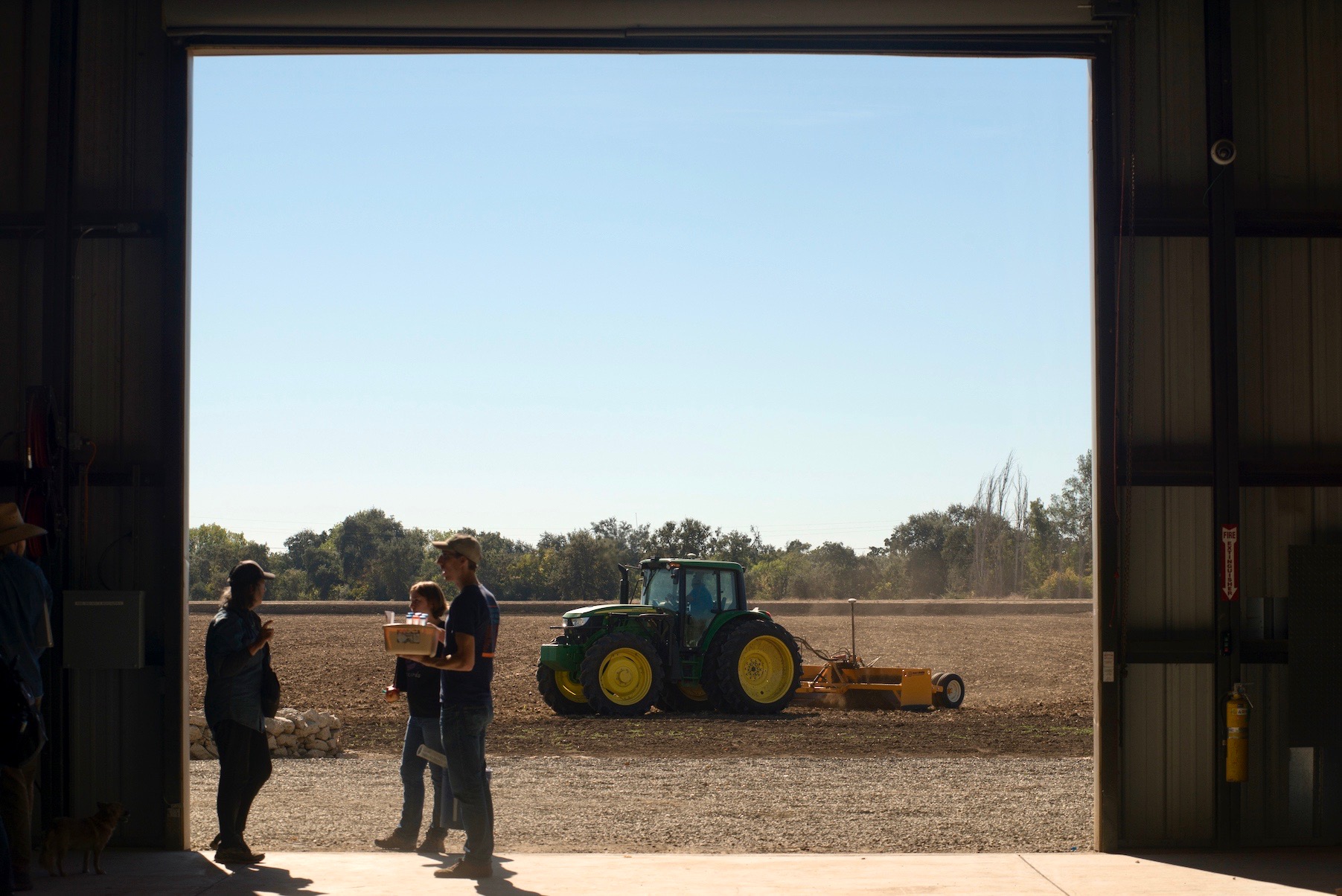 Woman talks with two students at UC Davis within frame of barn looking out on field of soil, with trees in distance