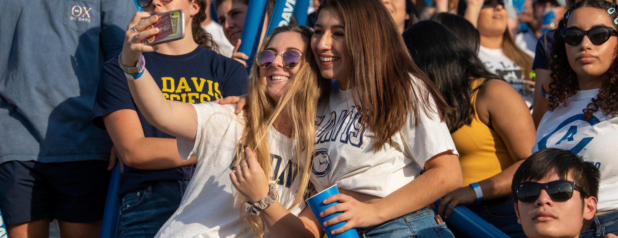 Two females students take a selfy in the crowd at a raucous football game