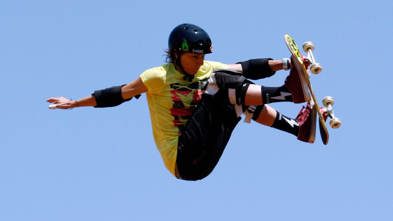 Photo of a woman on a skateboard doing a trick. She is above the level of the camera and completely surrounded by blue sky. She has one hand and both feet on the board.