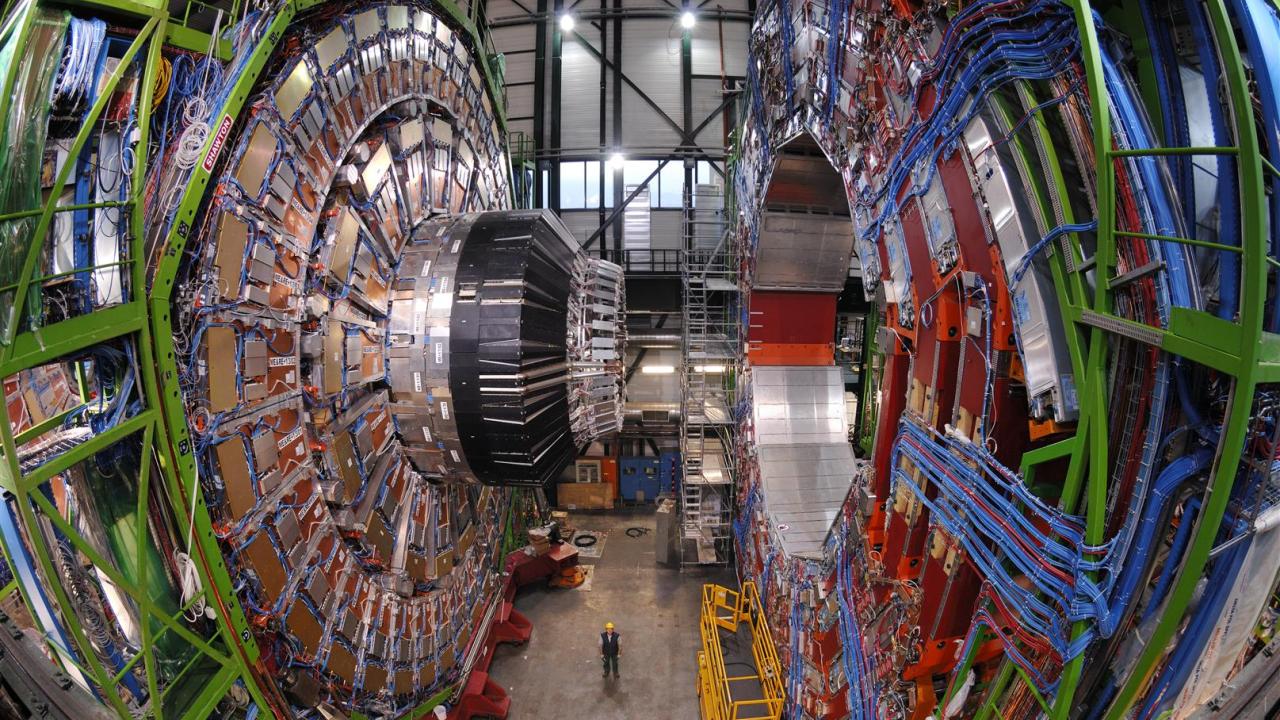 Large, complex machines with lots of colorful wiring dominate a hall with some small human figures visible. 