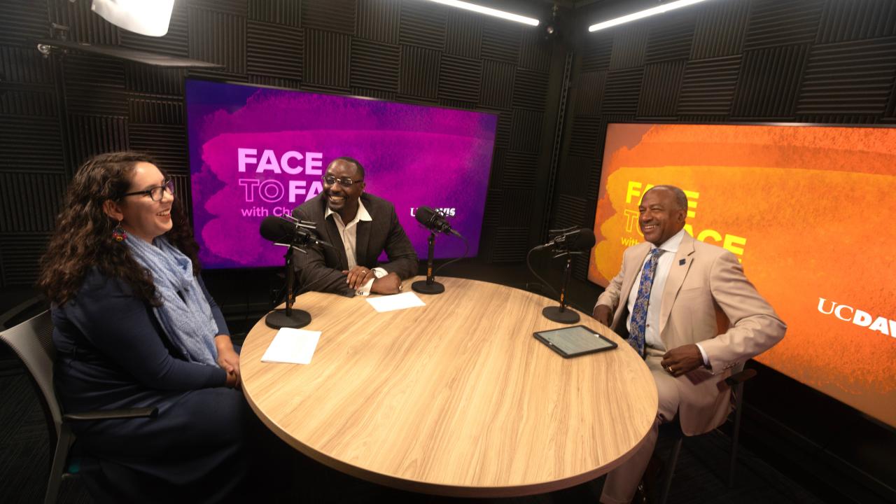 Inside a recording studio, three individuals sit at a round table. From left to right, Stephanie Zarate, Kayton Carter and Chancellor Gary May are seated and laughing in conversation while recording this podcast episode. 