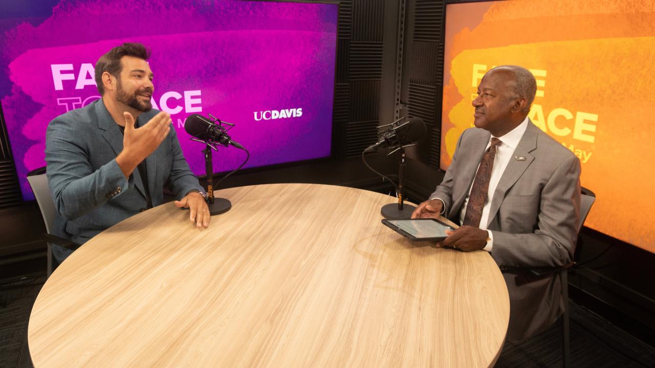 Two men sit across from each other at a round wooden table inside of a recording studio. The man on the left is wearing a dark teal suit jacket, has short dark hair and is speaking while moving his hands. On the right side, Chancellor Gary S. May wears a light brown suit and listen to Weber's talking points.