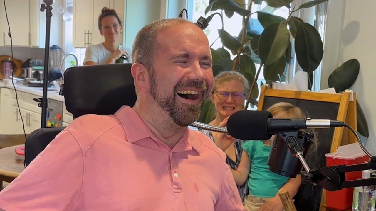 A man wearing a pink polo shirt weeps and laughs. In the background two other people can be seen weeping and smiling. 