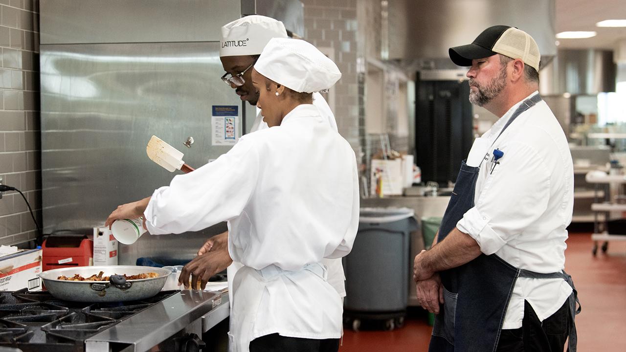 A chef oversees two people cooking at a dining facility stove.