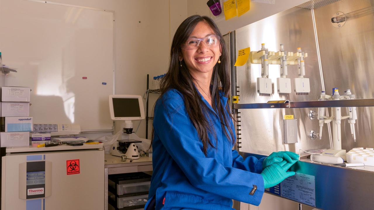 A smiling scientist in a blue lab coat working in a laboratory setting, with safety goggles on and handling lab equipment.