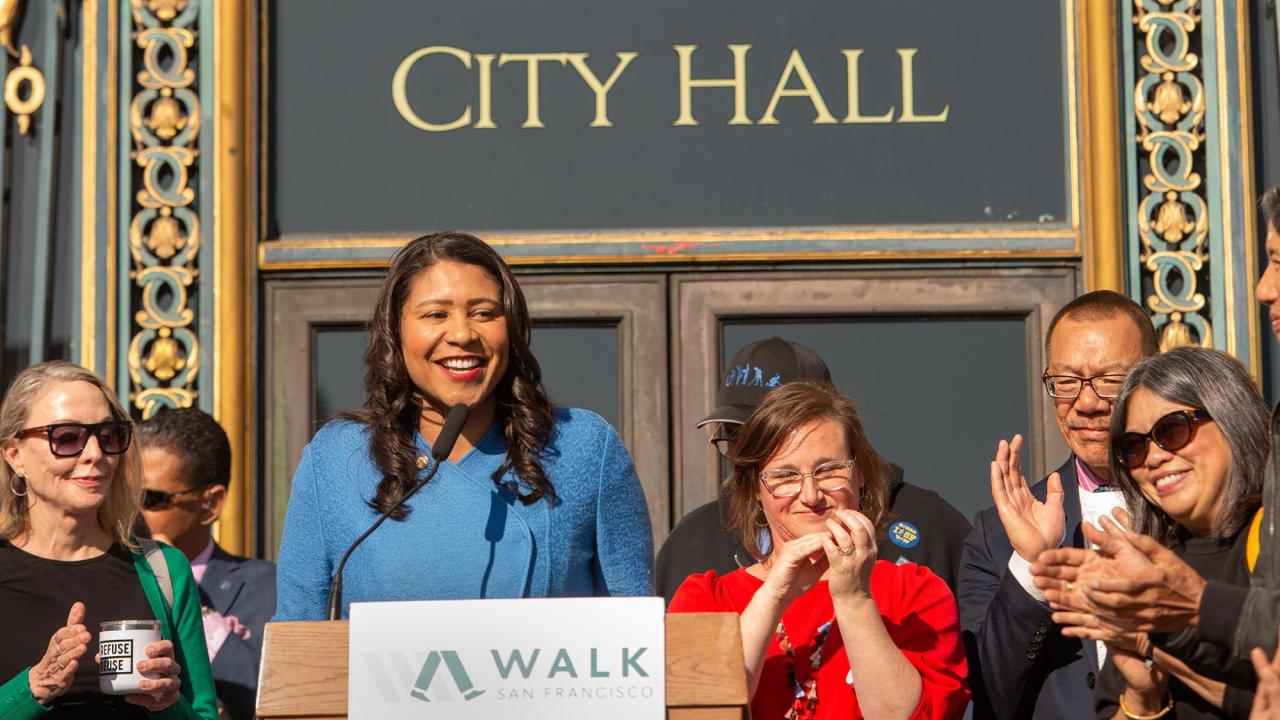 A woman stands at a podium in front of city hall while people listen.