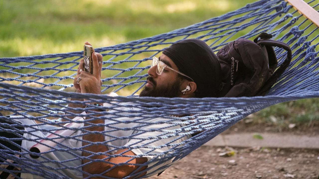 A male student lies in a blue hammock and holds his phone up to look at it.