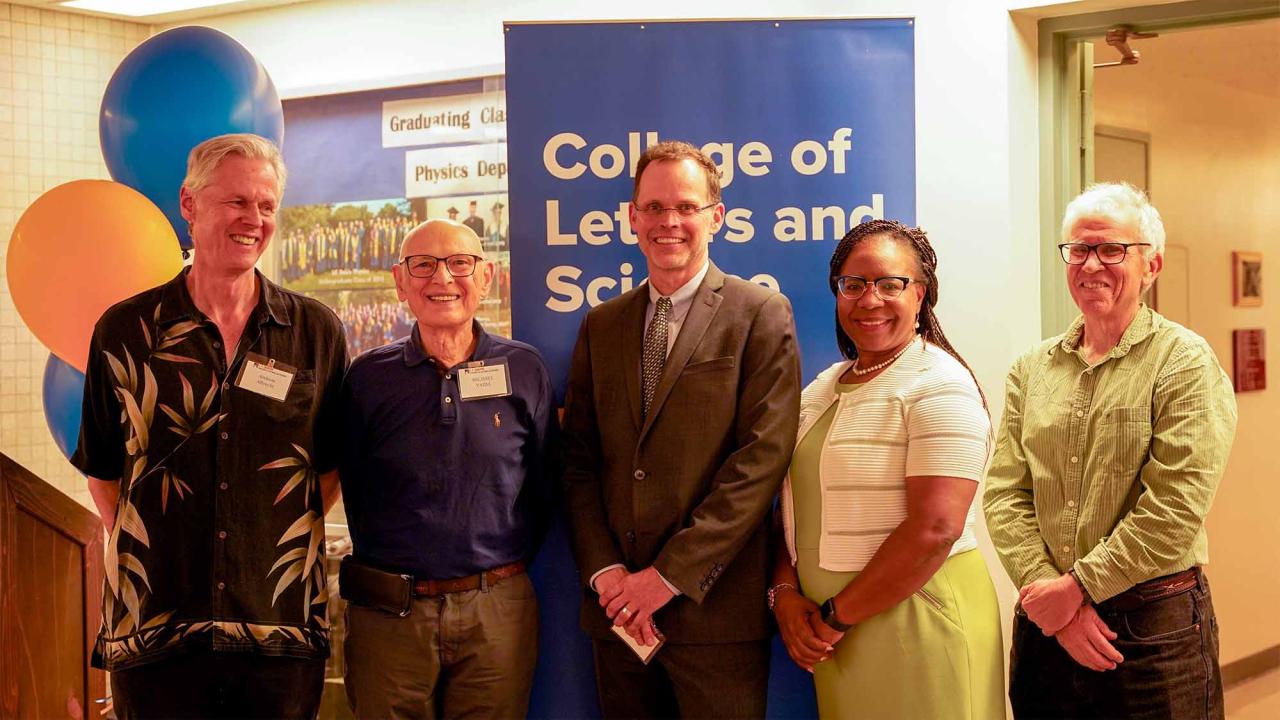 Five people stand smiling in front of a "College of Letters and Science" banner. From left: a tall man in a patterned shirt, an older man in a blue polo, a man in a suit and tie, a Black woman in a white top, and an older man in a yellow shirt. Colorful balloons are visible in the background.