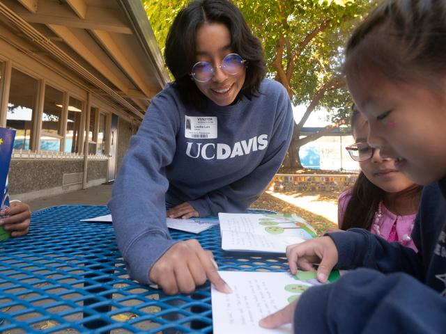 A woman points to the page of a book as two children look on