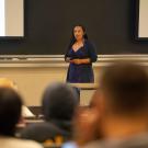 A professor teaches in front of a large lecture with a slide presentation behind them. 