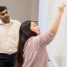 Business school student YiYi Han (foreground) at the white board writing while an instructor watches from behind.