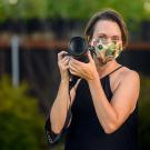 Woman holding camera, wearing face covering.