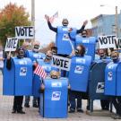 Theatre troupe in mailbox costumes, carrying "Vote" signs.