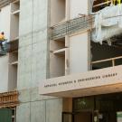 Workers in lift, on exterior of concrete building, with library's sign and front door in foreground.