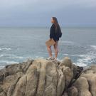 A woman stands on giant shoreline rocks looking out at the ocean and holding a clipboard