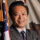 Portrait of a man wearing a gray suit and standing in front of the U.S. and California flags, with his arms crossed and smiling.