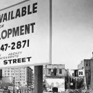 Black and white photo showing a large sign put up in front of a vacant lot. The sign reads "Land Available for Redevelopment" and gives a phone number and address. Buildings toward the back of the lot are undergoing various stages of demolition.