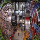 Large, complex machines with lots of colorful wiring dominate a hall with some small human figures visible. 