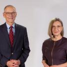 On the left, a white male with grey hair and glasses wearing a dark suit and red tie. On the right is a white woman with blonde hair and glasses wearing a deep purple blouse. 