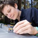 A woman in a dark blue shirt with short brown hair leans over a table outdoors. By her hand is a plastic tube with an insect inside. 