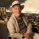 Inside an office sits a man in a straw fedora and beige jacket, smiling at the camera from a swivel chair in front of his desk, which has several different toy cars and other trinkets related to transportation. 