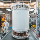 A large white cylinder in the middle of a laboratory setting. To the left a person in a white clean room coverall and mask descends a staircase. 