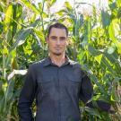 Head and shoulders of a man wearing a dark blue shirt standing in a cornfield. 