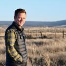 A man in a puffer vest and plaid short stands on a remote ranch, looking at the camera. Brushy fields and distant mountains are in the background.