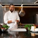 A male chef in a white coat puts white rubber gloves on his hands. He is standing in a kitchen behind a black countertop that has a white chopping board, raw meat and vegetables on it.