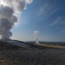 On the left, a large cloud of vapor rising from the ground with another geyser erupting in the background, against a blue sky. 