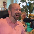 A man wearing a pink polo shirt weeps and laughs. In the background two other people can be seen weeping and smiling. 