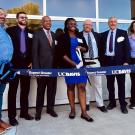 A group of men and women in semi-formal dress stand behind a blue ribbon held by stanchions. In the center, a black woman is about to cut the ribbon with large scissors. 