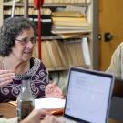 Professor of Anthropology explaining her work to a student at a table with computers and tablets.