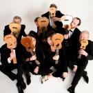 Eight orchestra members sit against a white background and hold a ukulele next to their face.