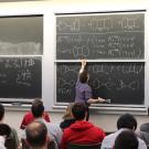 From the rear of a classroom, rows of students face a blackboard where a professor is drawing symbols. 