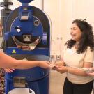 Three people stand around a large blue coffee roasting machine. On the left, a blonde woman in a patterned sleeveless top is handing something to a curly-haired woman in the middle, who is wearing a light-colored shirt. On the right stands a man with long hair tied back, wearing a light gray t-shirt. All three are smiling and engaged in conversation. The prominent blue coffee roaster bears the "Probat" brand name.