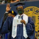 Graduate in commencement regalia (and mask) waves as he walks across stage