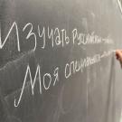 A hand writes on a blackboard in white chalk. The words are Russian, printed in Cyrillic.