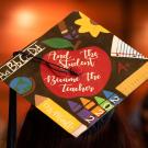 A grad cap that reads, "And the student became the teacher," in white cursive within a bright red apple, surrounded by teaching equipment like rulers, crayons and a pencil that reads, "Ms. Murillo." The photo was taken at UC Davis' School of Education Commencement graduation ceremony in 2023.