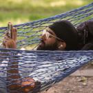 A male student lies in a blue hammock and holds his phone up to look at it.