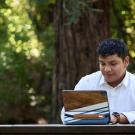 A student in a white button-down shirt, works on their iPad outside at a table in the UC Davis Redwood Grove.  